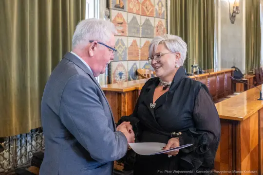 Uścisk dłoni dwóch osób w eleganckich strojach w sali urzędowej.