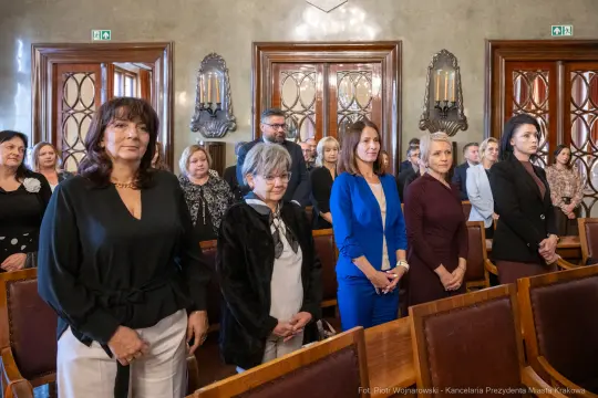 Grupa kobiet w eleganckich strojach, stojących w sali z dekoracyjnymi detalami.