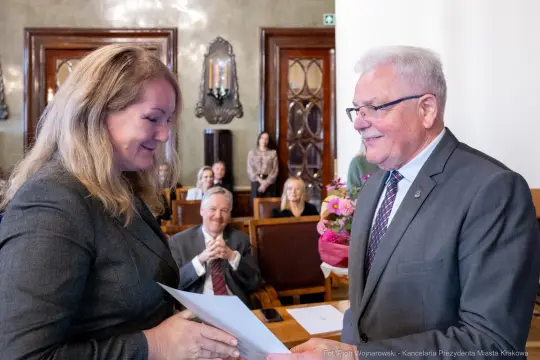 Wręczenie nagrody w sali obrad, uśmiechnięte twarze, kwiaty na stole.