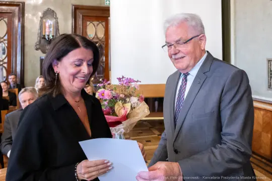 Wręczenie dyplomu z bukietem kwiatów w tle, uśmiechnięte twarze uczestników.