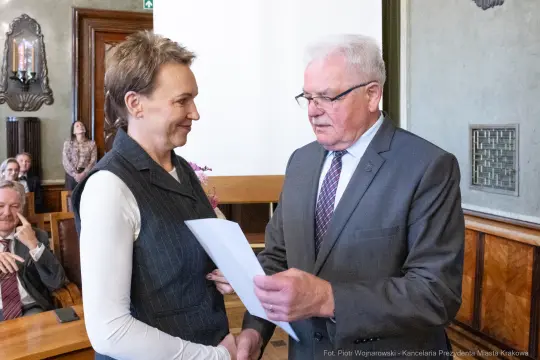 Wręczenie dokumentu w sali obrad, dwie osoby w formalnych strojach, w tle zebrani goście.