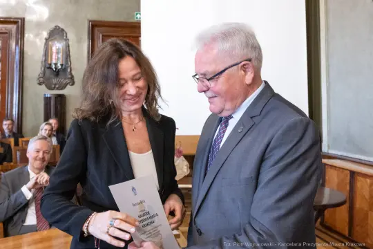 Wręczenie dyplomu przez mężczyznę kobiecie w eleganckim otoczeniu. W tle widoczni inni uczestnicy.