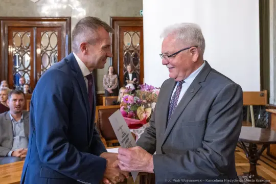 Uroczystość wręczenia nagrody, uściski dłoni, kwiaty w tle.