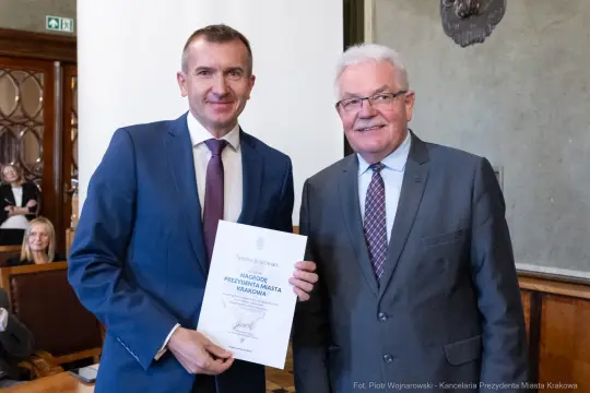 Dwaj mężczyźni trzymają dyplom w sali urzędowej.