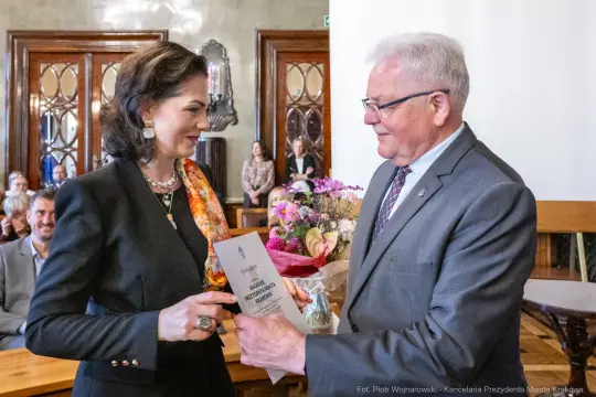 Wręczenie nagrody z bukietem kwiatów w tle, uśmiechnięte osoby w eleganckich strojach.