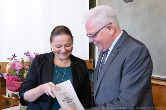 Uśmiechnięte osoby wręczające sobie dokument w otoczeniu kwiatów.