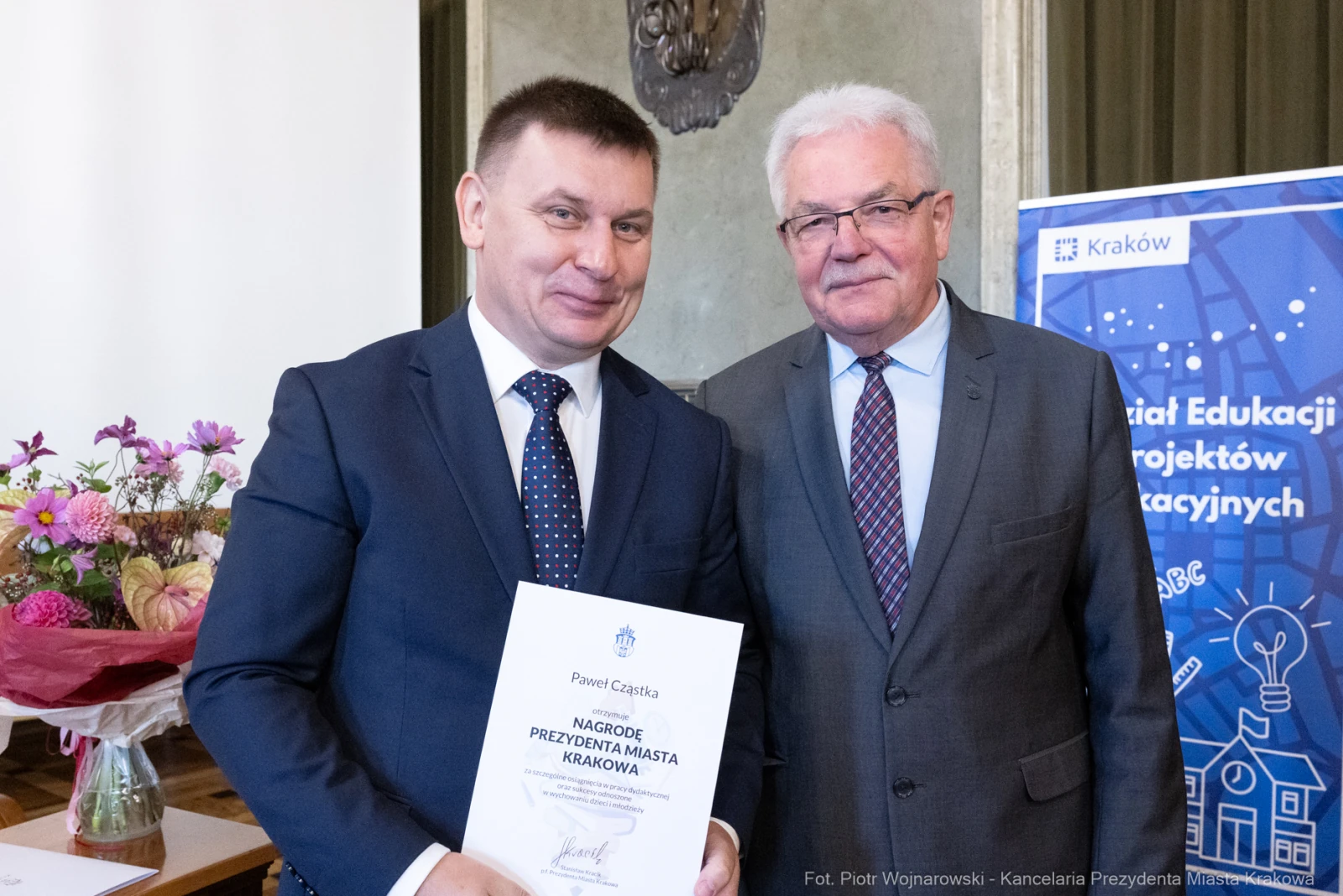 Dwóch mężczyzn trzyma dyplom nagrody w sali z kwiatami w tle.  Autor: Piotr Wojnarowski