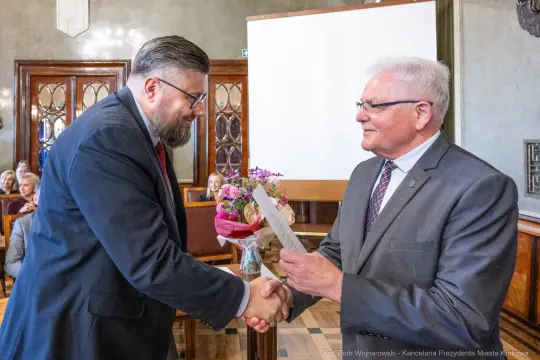 Wręczenie dokumentu podczas uroczystości, mężczyźni uścisną ręce, w tle kwiaty i publiczność.