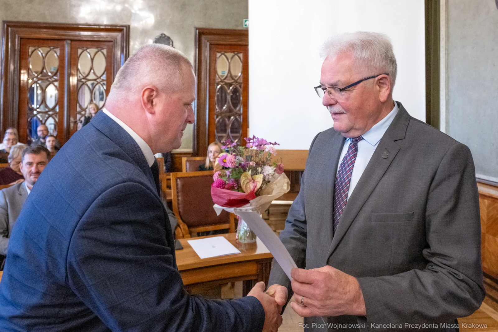Mężczyzna wręcza kwiaty i dokument innemu mężczyźnie w sali obrad.  Autor: Piotr Wojnarowski