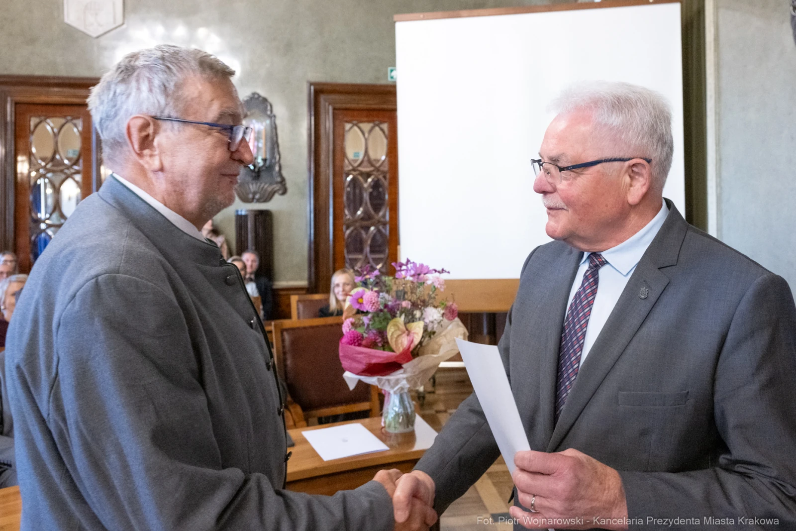 Dwaj mężczyźni uścisnęli sobie dłonie podczas ceremonii wręczenia nagrody. W tle kwiaty i publiczność.  Autor: Piotr Wojnarowski