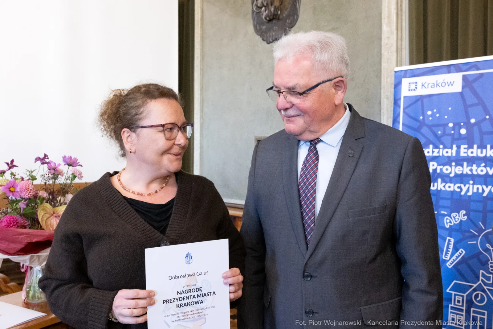 Kobieta z dyplomem i mężczyzna w garniturze, oboje uśmiechnięci, w tle dekoracje i baner.  Autor: Piotr Wojnarowski