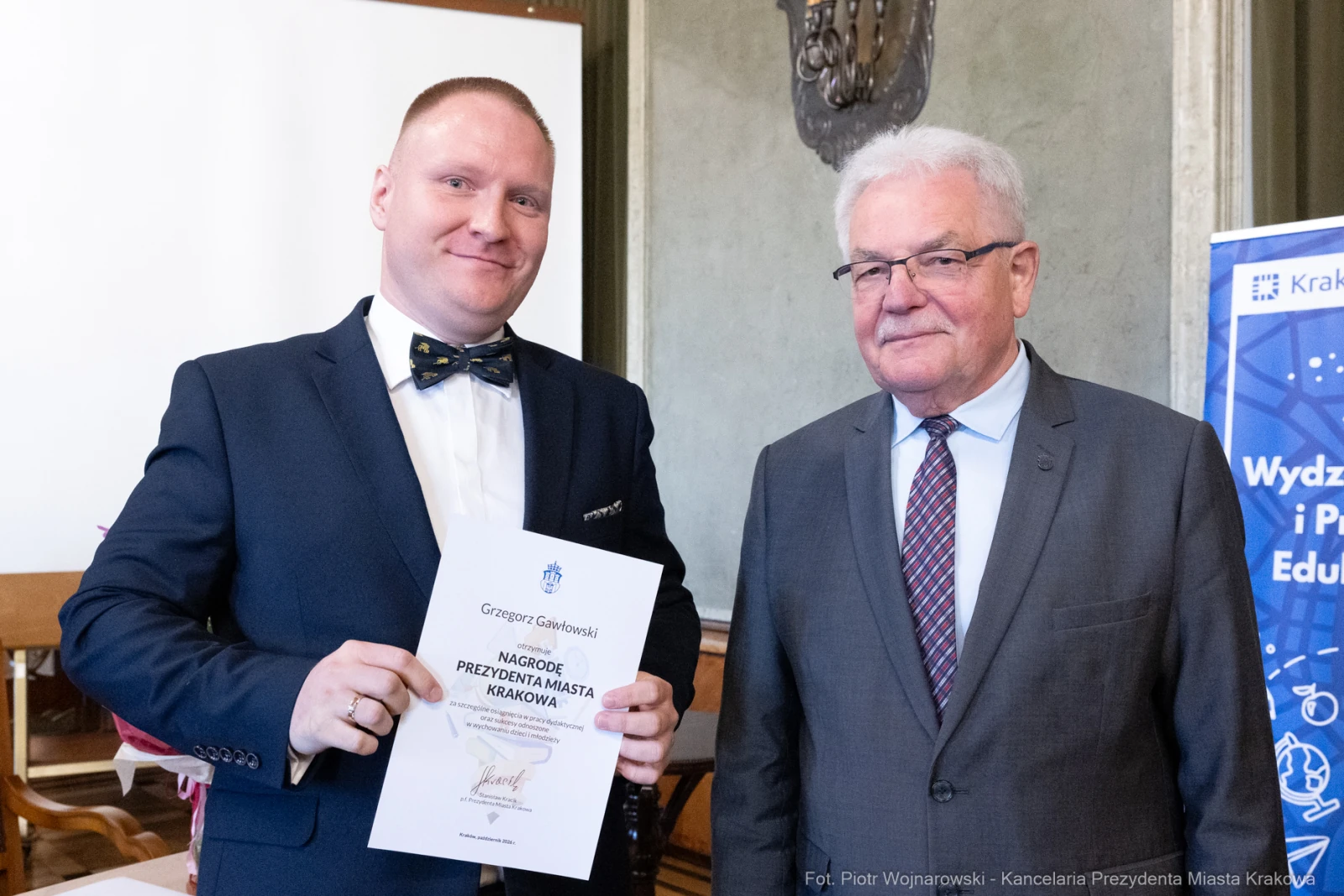 Mężczyzna w garniturze trzyma dyplom, obok stoi starszy mężczyzna w okularach.  Autor: Piotr Wojnarowski