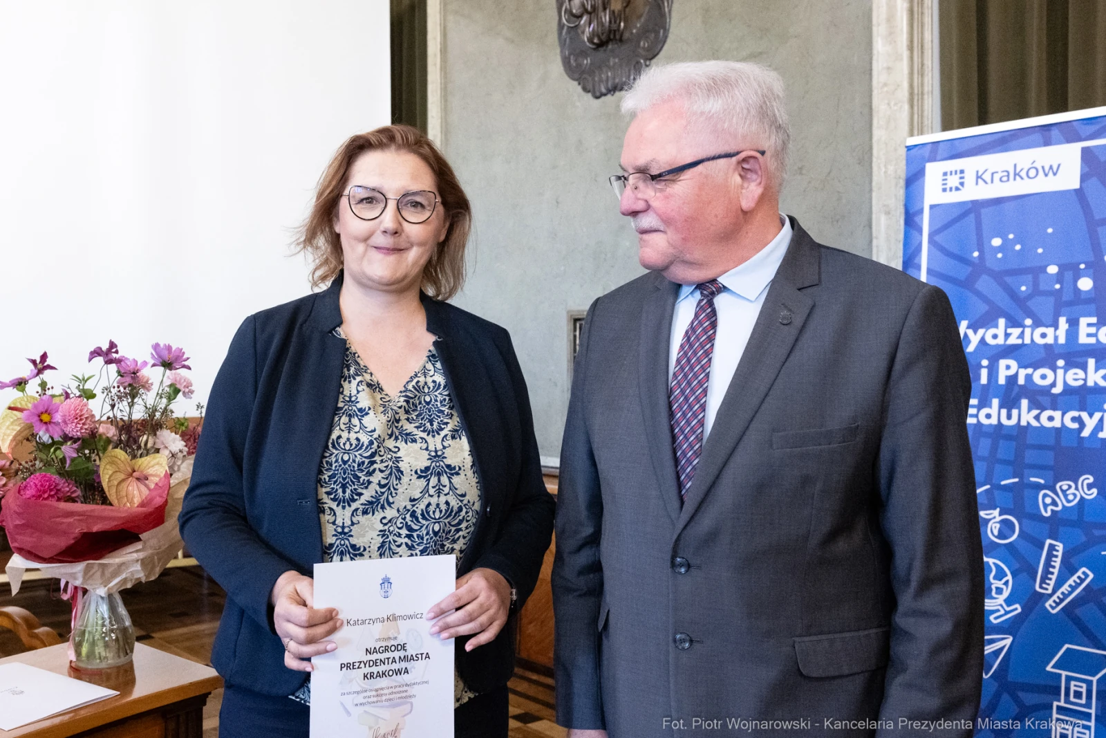 Kobieta z dyplomem i mężczyzna w garniturze, w tle kwiaty i baner informacyjny.  Autor: Piotr Wojnarowski