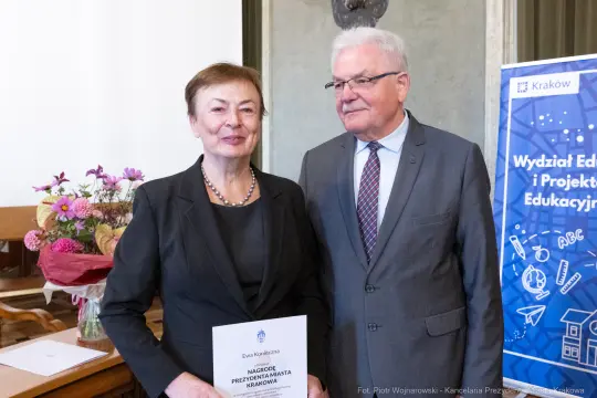 Kobieta z dyplomem i mężczyzna w garniturze, oboje uśmiechnięci, w tle baner edukacyjny.