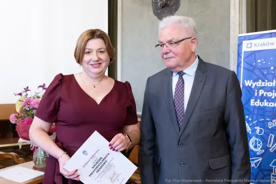 Kobieta z dyplomem i mężczyzna w garniturze, w tle dekoracje i logo uczelni.