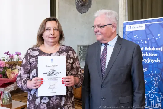 Kobieta trzymająca dyplom, obok niej mężczyzna w okularach. Uroczystość wręczenia nagrody.
