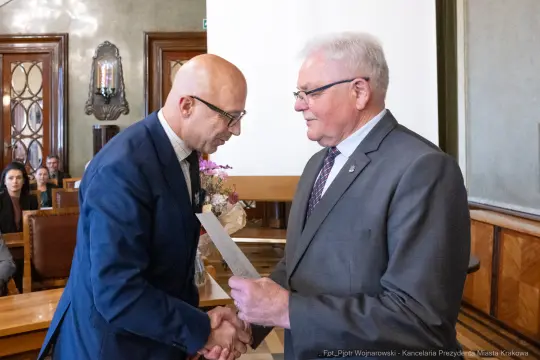 Wręczenie dyplomu w sali urzędowej, dwóch mężczyzn w formalnych strojach, kwiaty na stole.