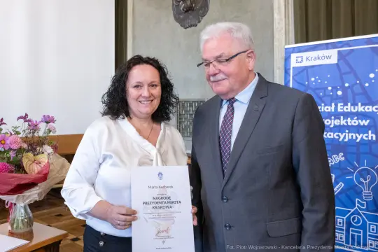 Kobieta odbierająca nagrodę od mężczyzny, w tle kwiaty i baner promujący edukację.