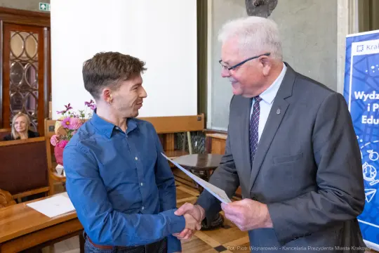 Mężczyzna odbierający dyplom od drugiego mężczyzny w formalnym otoczeniu.