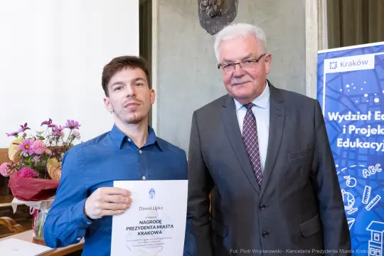 Mężczyzna trzyma dyplom, obok stoi starszy mężczyzna w okularach.