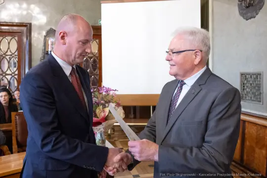 Mężczyźni w garniturach wymieniają uścisk dłoni, jeden z nich wręcza dokument. W tle kwiaty i publiczność.