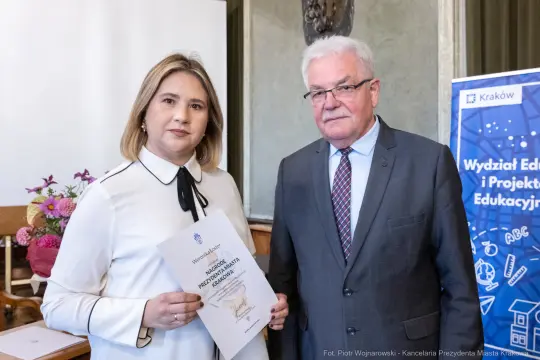 Wręczenie dyplomu przez mężczyznę kobiecie w sali z dekoracjami.