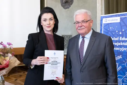 Kobieta trzymająca dyplom, obok niej mężczyzna w garniturze, w tle dekoracje i kwiaty.