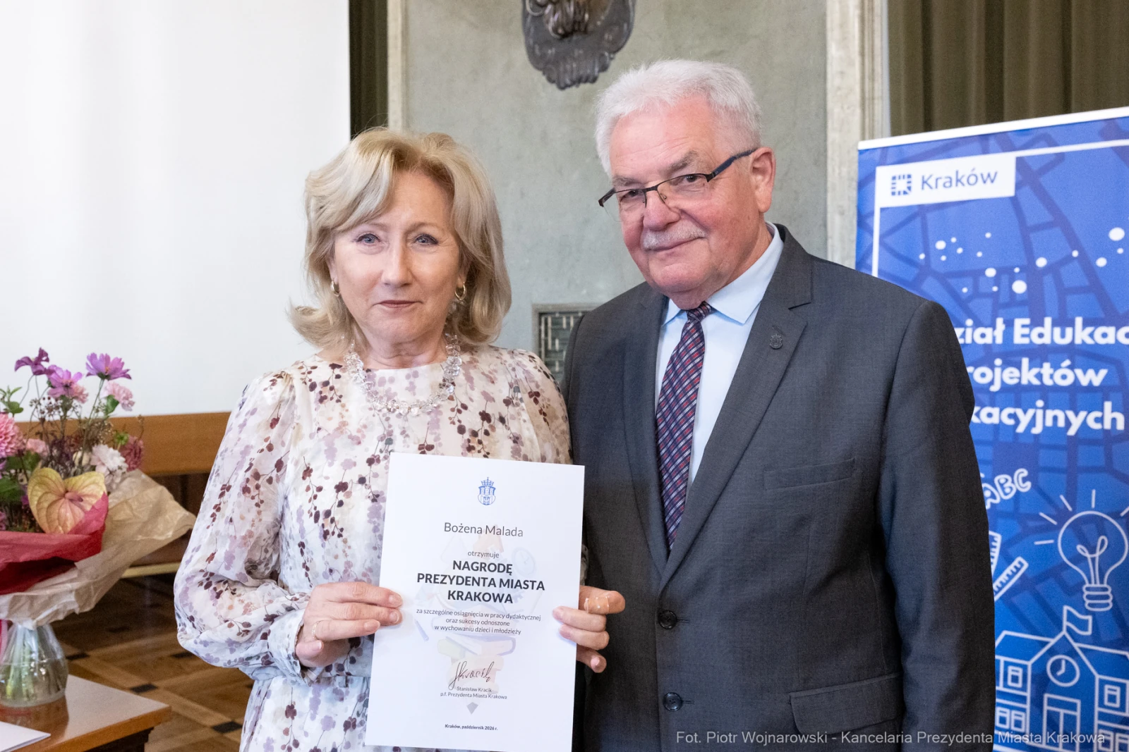 Kobieta odbierająca nagrodę od mężczyzny, w tle kwiaty i baner edukacyjny.  Autor: Piotr Wojnarowski