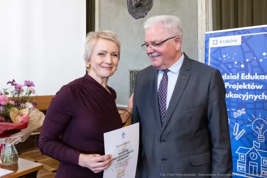 Kobieta wręcza dyplom mężczyźnie w formalnym otoczeniu, obok kwiaty i plakat edukacyjny.
