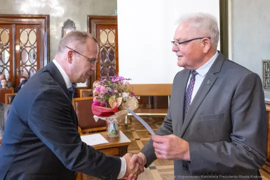 Mężczyźni w garniturach wymieniają uściski dłoni, jeden z nich trzyma bukiet kwiatów.