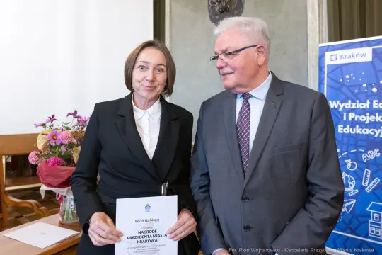 Kobieta w eleganckim stroju trzyma dyplom, obok mężczyzna w garniturze. Tło z logo Krakowa.