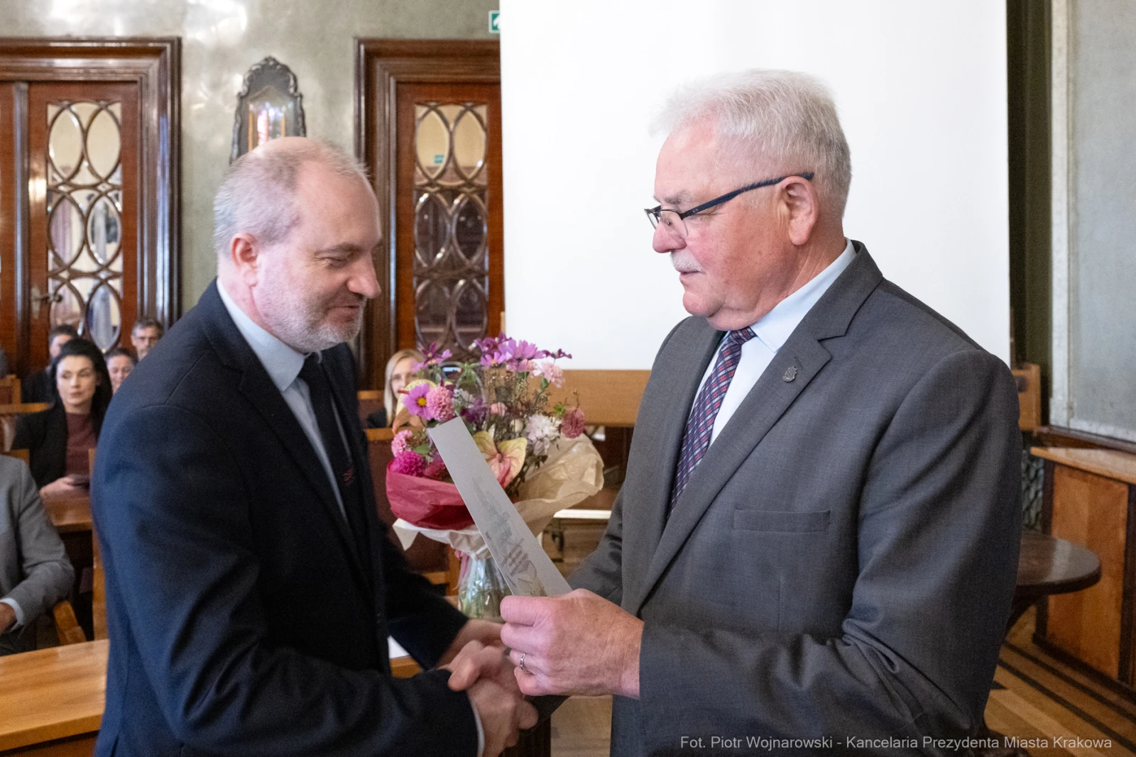 Mężczyźni w formalnych strojach, jeden wręcza dokument, drugi trzyma bukiet kwiatów.  Autor: Piotr Wojnarowski