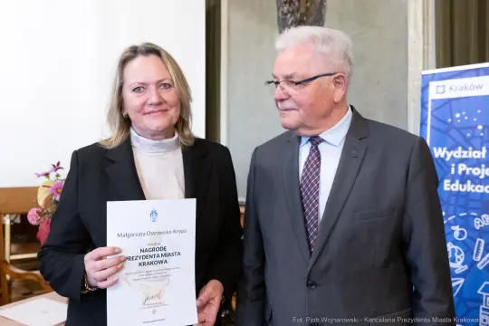 Kobieta trzyma dyplom, obok niej stoi mężczyzna w okularach. W tle widoczny banner z logo Krakowa.
