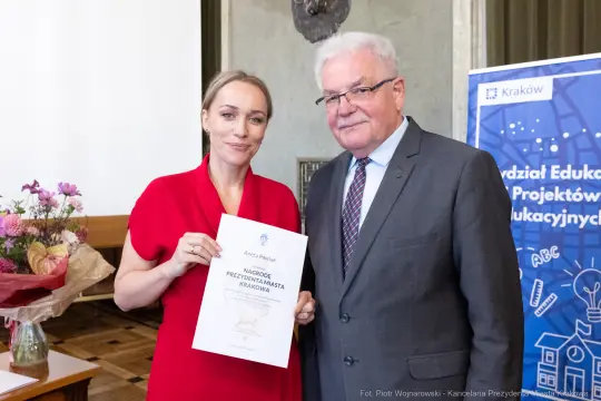 Kobieta i mężczyzna trzymają dyplom nagrody, w tle kwiaty i baner edukacyjny.