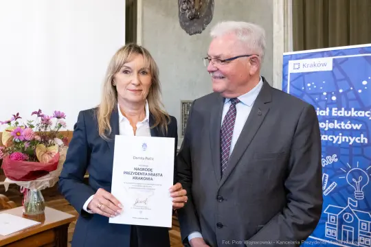 Dorota Pelc odbiera nagrodę od przedstawiciela miasta Krakowa. W tle kwiaty i dekoracje sali.