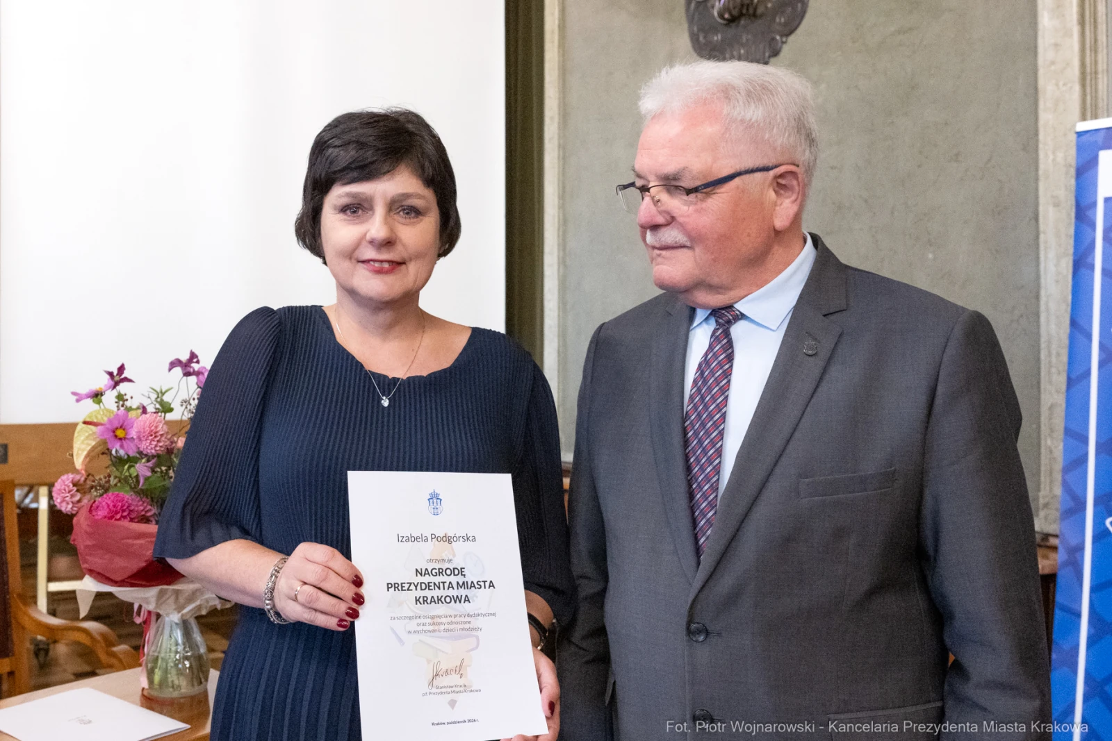 Kobieta trzymająca dyplom, obok mężczyzna w garniturze, w tle kwiaty i stół.  Autor: Piotr Wojnarowski
