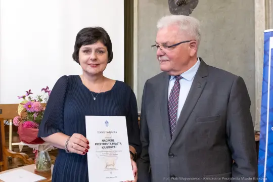 Kobieta trzymająca dyplom, obok mężczyzna w garniturze, w tle kwiaty i stół.