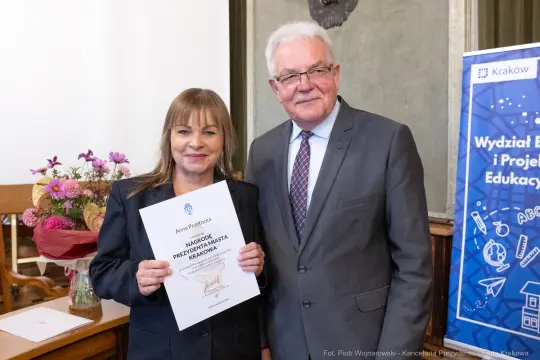 Wręczenie nagrody dla osoby z dyplomem w towarzystwie mężczyzny w garniturze.