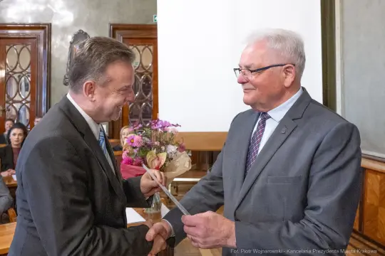 Dwaj mężczyźni uścisnęli sobie dłonie, jeden wręcza drugi dokument, w tle kwiaty i osoby siedzące.