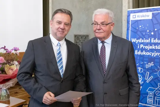 Dwaj mężczyźni w garniturach, jeden trzyma dokument, obaj uśmiechają się. W tle baner edukacyjny.