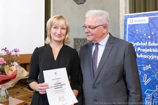 Kobieta z dyplomem i mężczyzna w garniturze, oboje uśmiechnięci, w tle kwiaty i grafika edukacyjna.