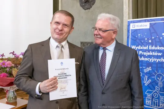 Mężczyzna z dyplomem obok drugiego mężczyzny, uśmiechają się w formalnym otoczeniu.