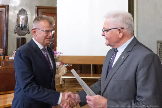 Dwaj mężczyźni uścisnęli sobie dłonie, jeden wręcza dokument drugiemu. W tle kwiaty i sala konferencyjna.