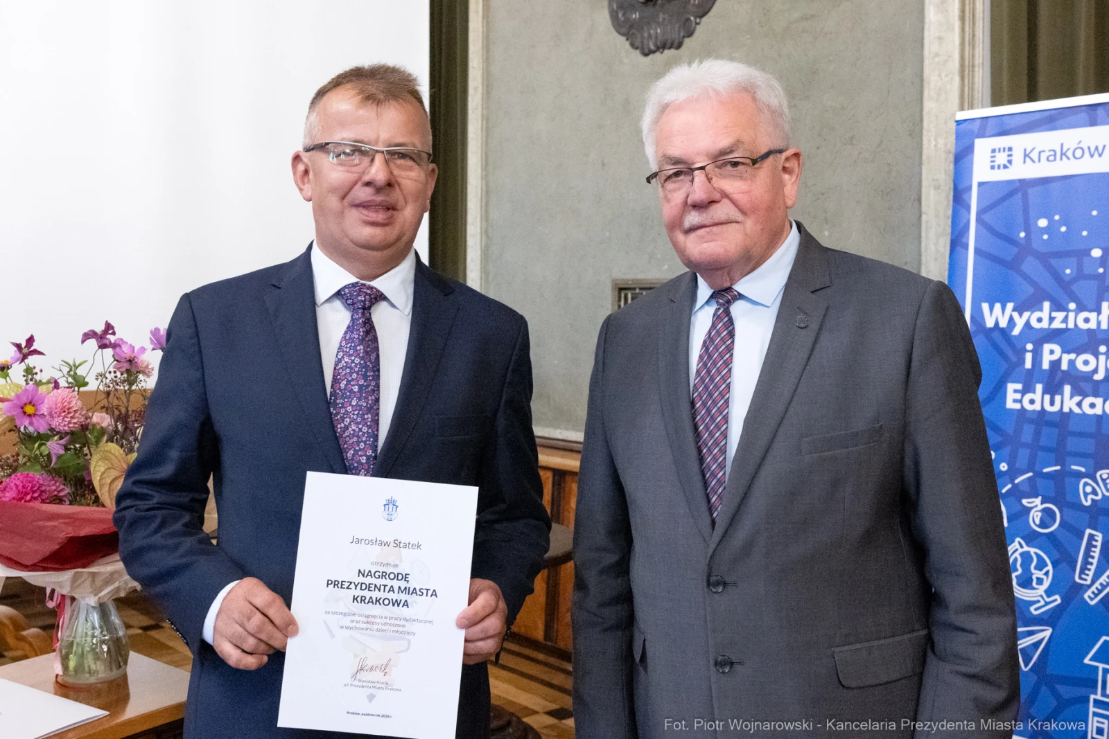 Dwaj mężczyźni trzymają dyplom w sali z kwiatami i banerem promującym edukację.  Autor: Piotr Wojnarowski