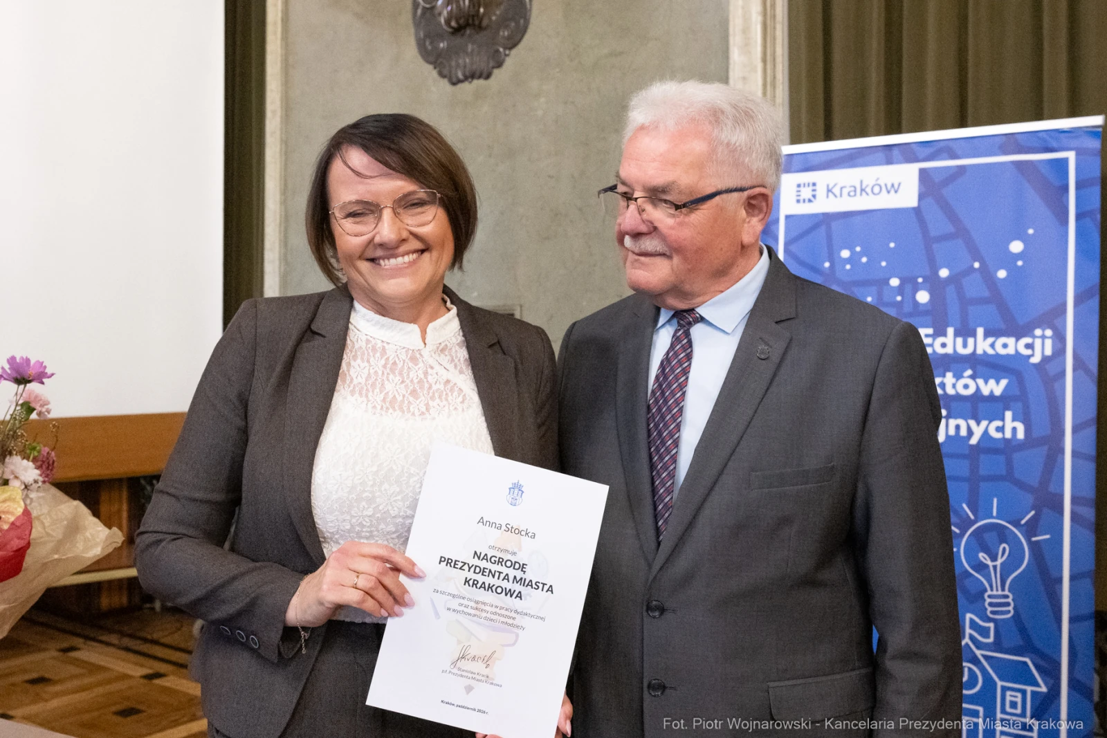 Kobieta trzymająca dyplom, obok niej mężczyzna w garniturze, w tle dekoracje i logo Krakowa.  Autor: Piotr Wojnarowski