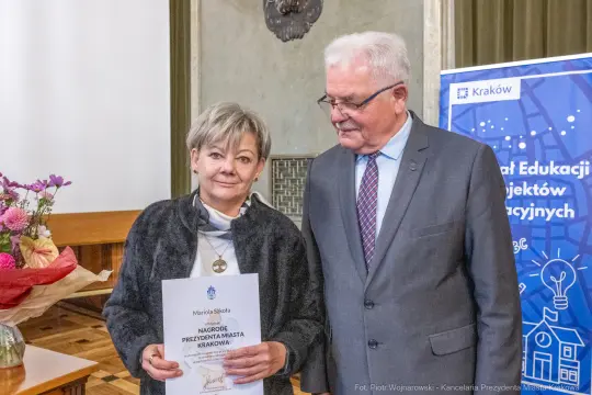 Kobieta z dyplomem i mężczyzna w garniturze, w tle kwiaty i elementy dekoracyjne.
