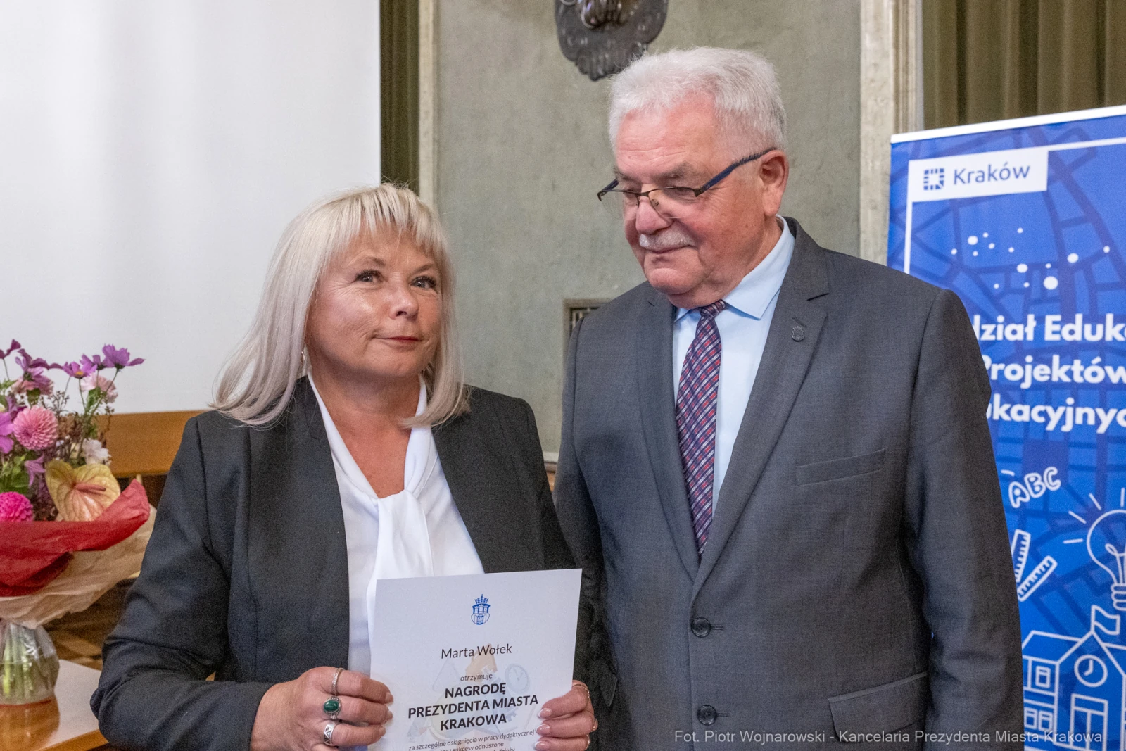 Kobieta odbierająca nagrodę od mężczyzny w formalnym stroju, w tle kwiaty i plakat edukacyjny.  Autor: Piotr Wojnarowski