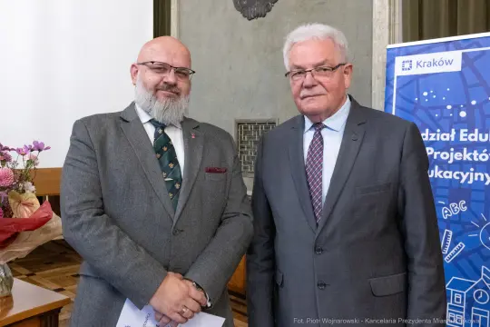 Dwóch mężczyzn w formalnych strojach, stojących obok siebie w sali.