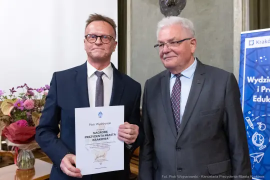 Dwóch mężczyzn trzyma dyplom nagrody Prezydenta Miasta Krakowa. W tle kwiaty i baner edukacyjny.