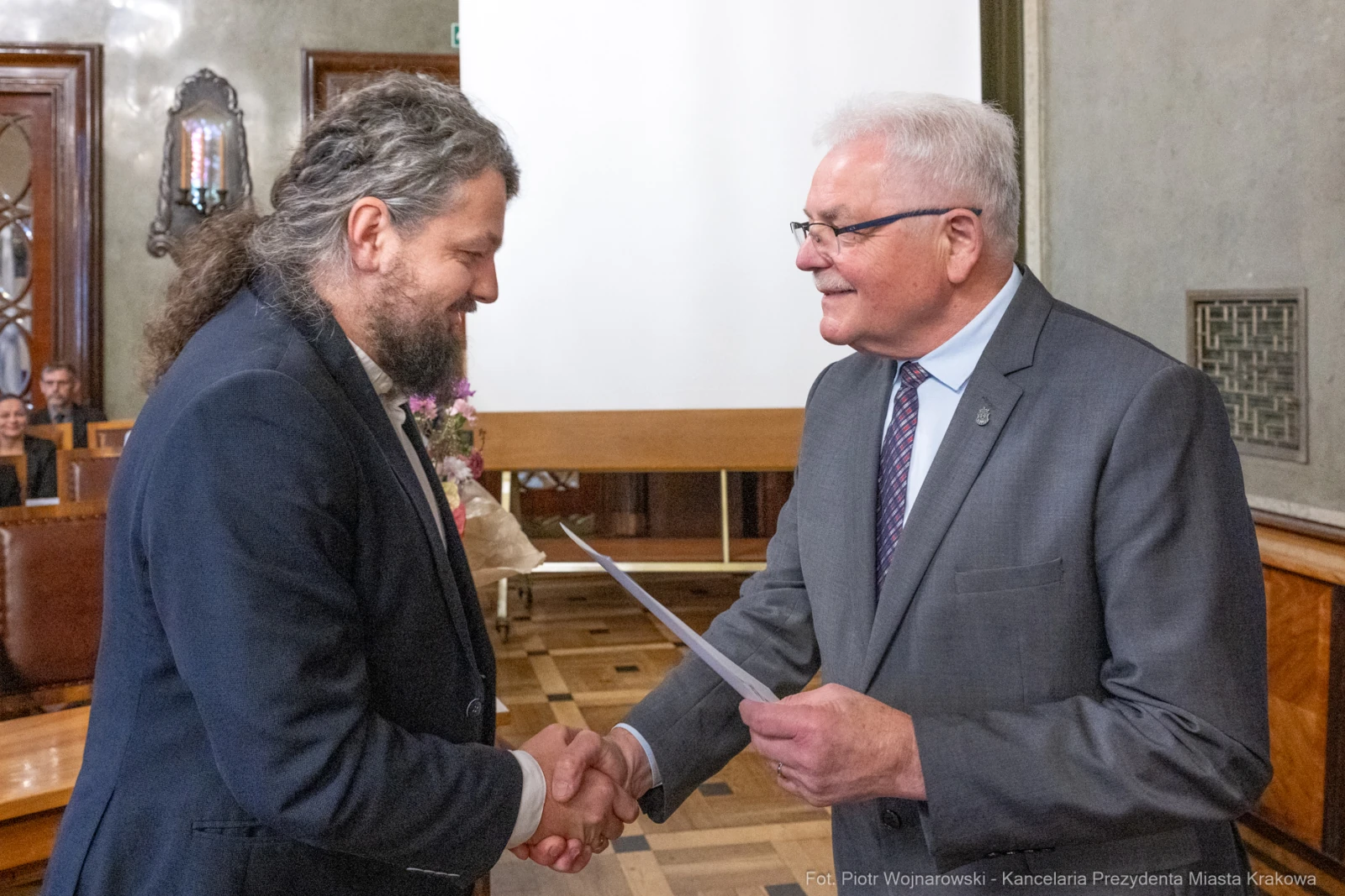 Mężczyźni uściskiem dłoni wymieniają dokumenty w formalnej atmosferze.  Autor: Piotr Wojnarowski